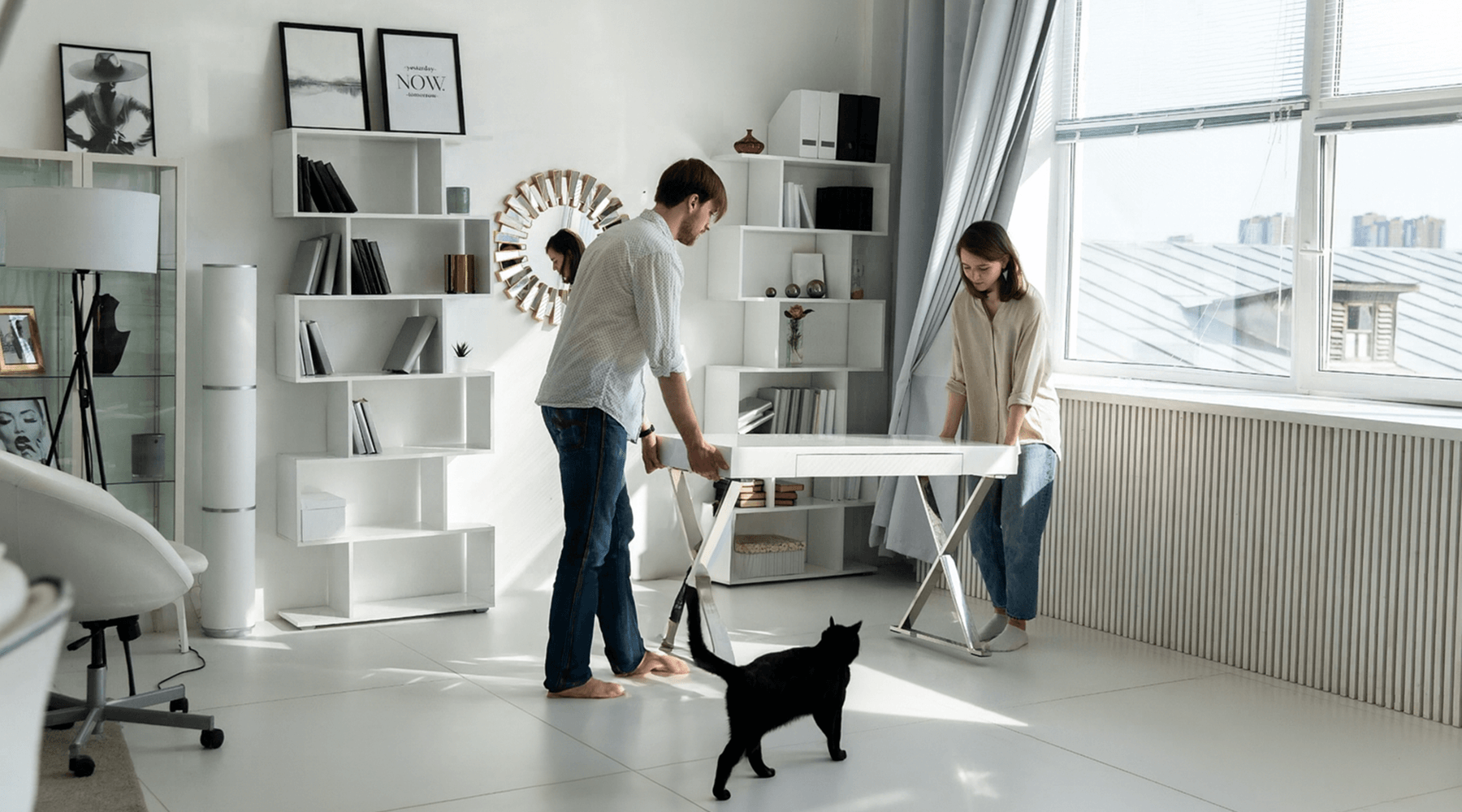 Couple rearranging furniture in a bright living room with a black cat walking nearby